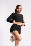 Confident woman in a black knit crop top and shorts, standing on one leg with a slight smile