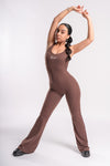 A woman dressed in a form-fitting brown jumpsuit poses with her hands on her hips.