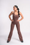 A woman in a brown fitted jumpsuit stands confidently, with hands on her hips, looking to the side