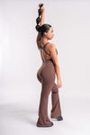 A woman with a long ponytail stands confidently in a brown jumpsuit, hands on hips, striking a pose.