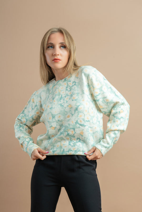 A woman in a pastel floral sweater and black pants poses with hands on her hips