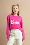 Woman wearing a bright pink sweatshirt with a white Barbie logo paired with off-white high-waisted pants