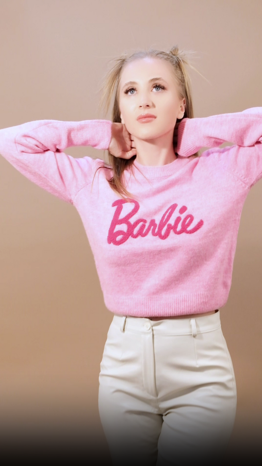 Woman wearing a baby pink pullover with a dark pink embroidered Barbie logo